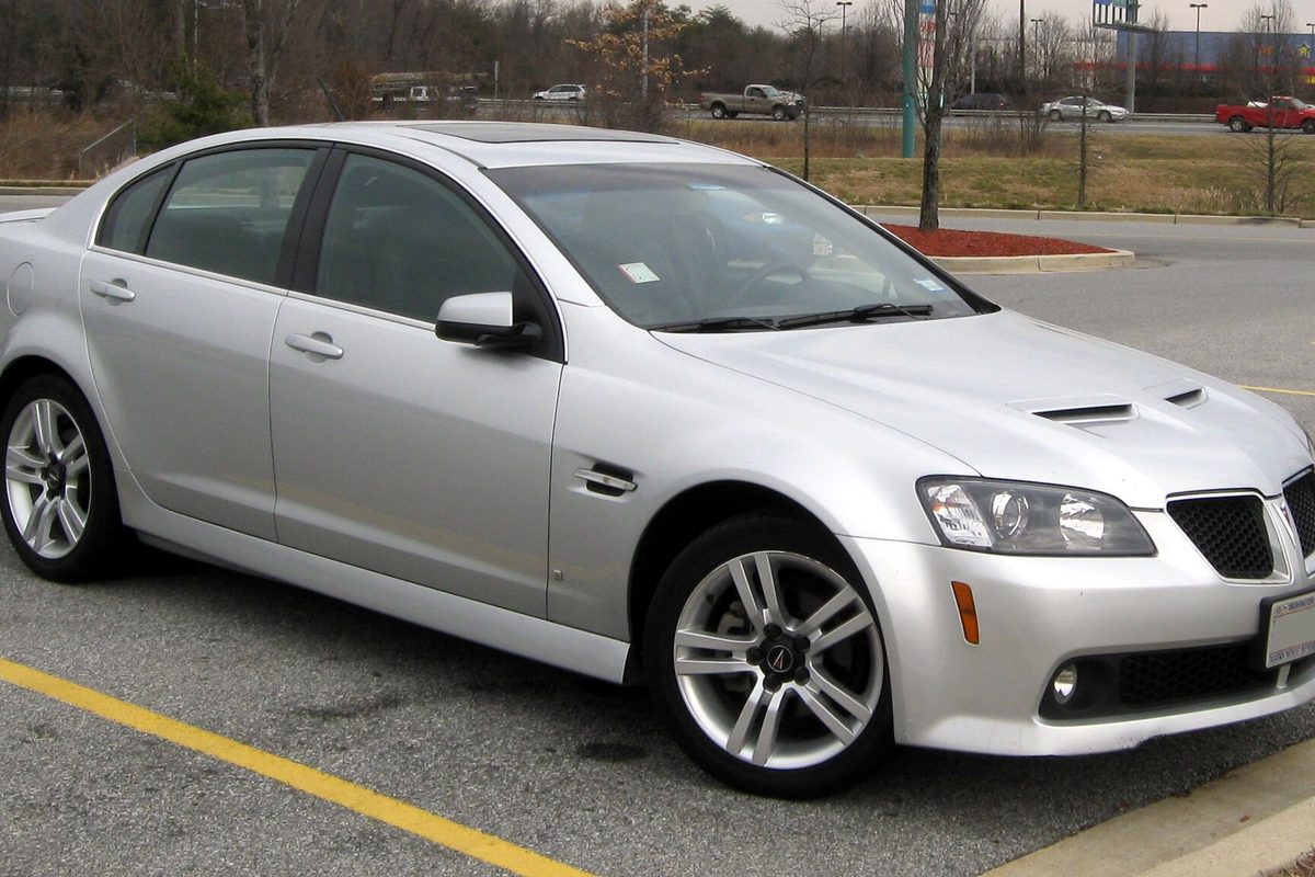 A silver Pontiac G8 sedan with twin hood scoops and five-spoke alloy wheels, parked beside a yellow curb line in a parking lot