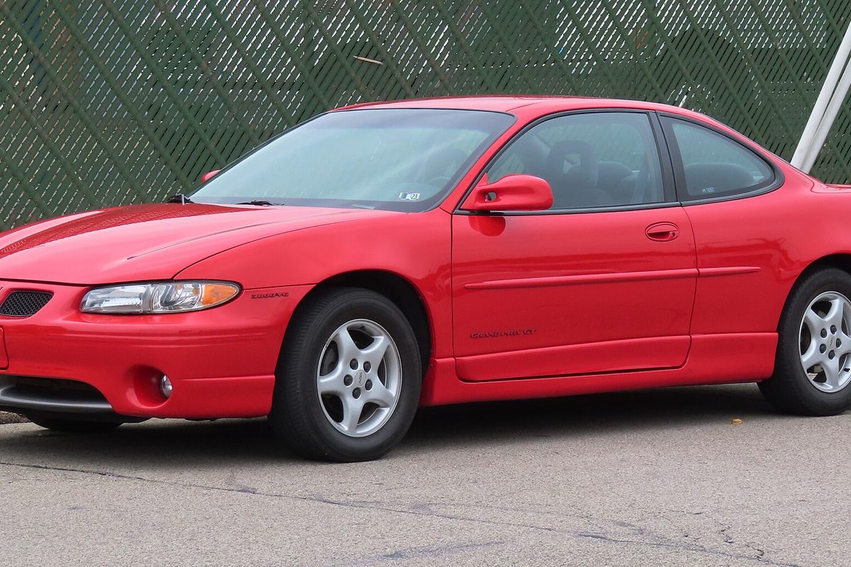 A bright red seventh-generation Pontiac Grand Prix GT coupe with silver alloy wheels, parked by a green fence