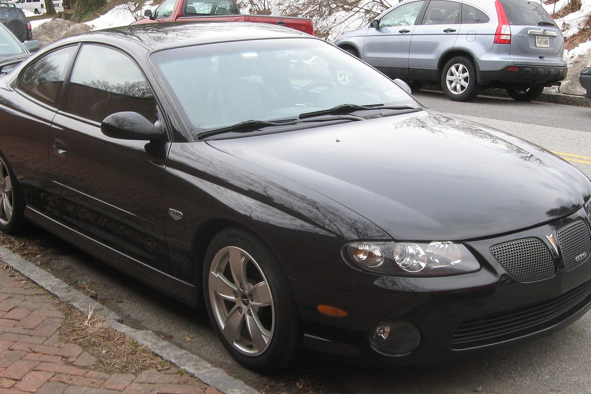A black Pontiac GTO coupe parked at a brick-edged curb, with snow on the ground behind it