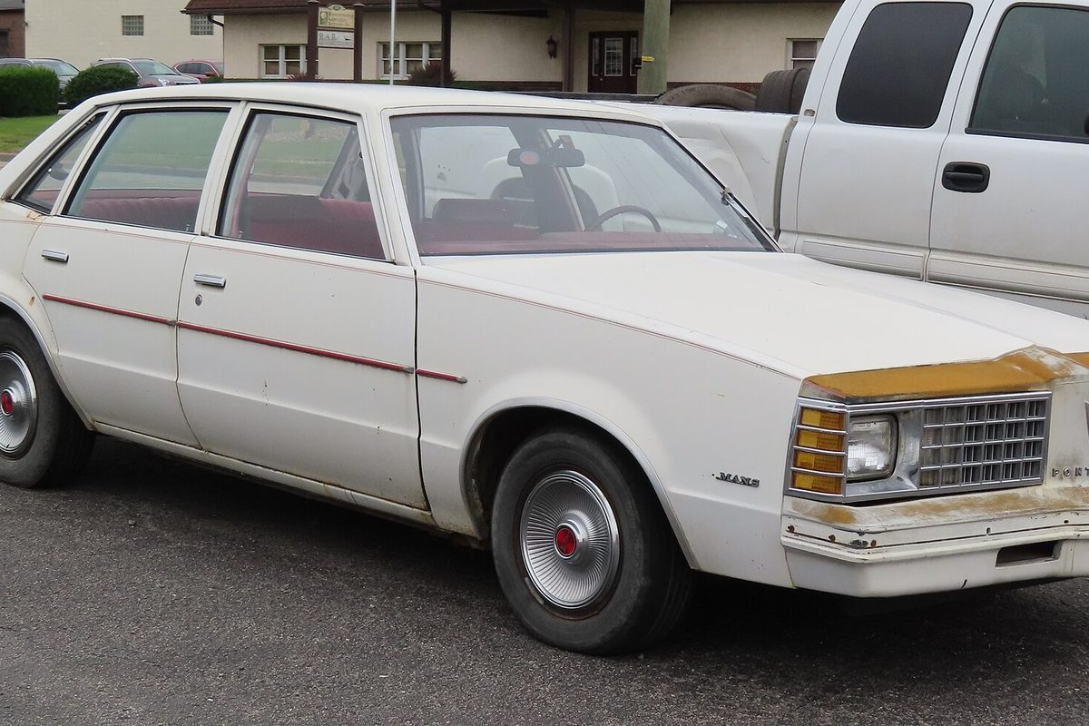 A white fifth-generation Pontiac LeMans four-door sedan with a red pinstripe, red interior and rust on the front bumper, parked beside a pickup