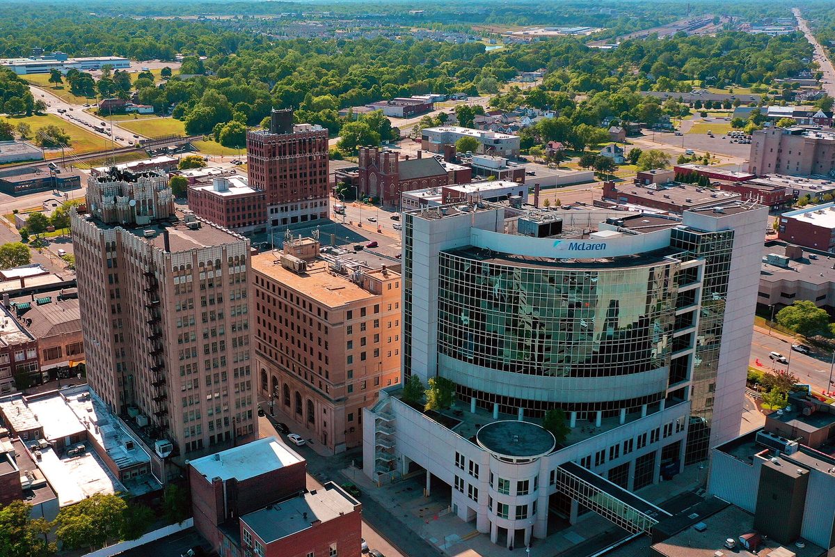 Aerial view of downtown Pontiac, older brick high-rises beside a curved glass medical building, with green trees stretching beyond