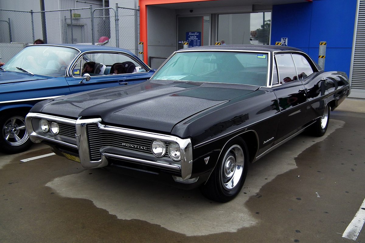 A black 1968 Pontiac Parisienne four-door hardtop with a chrome grille and quad headlights, parked on wet pavement at a car show