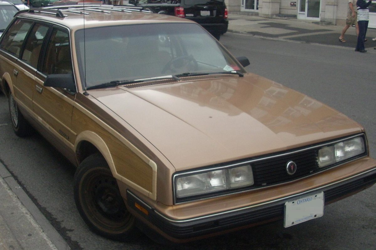 A tan Pontiac 6000 Safari station wagon with woodgrain side panels and a roof rack, parked on a city street