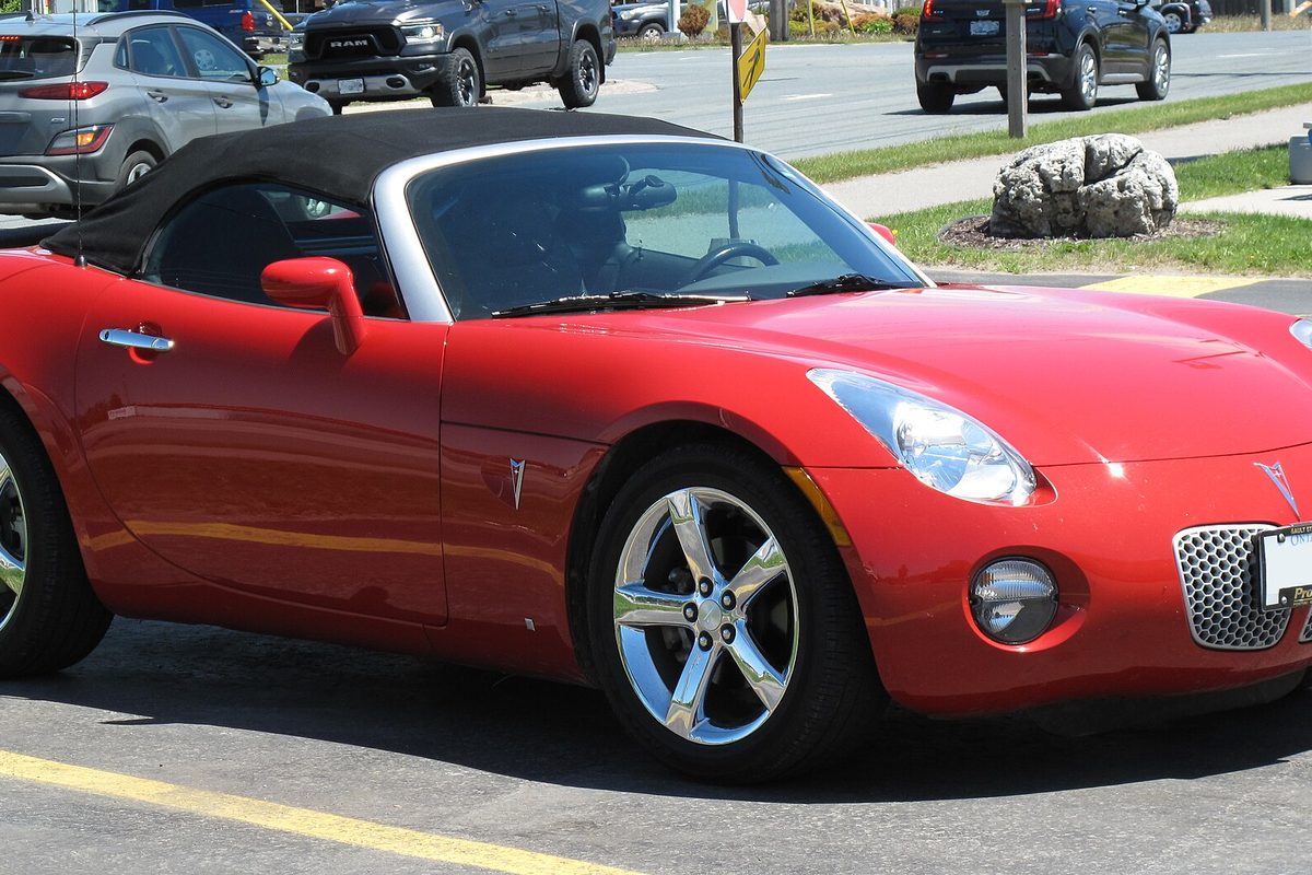 A red Pontiac Solstice roadster with its black soft top raised, parked in a lot beside a lawn