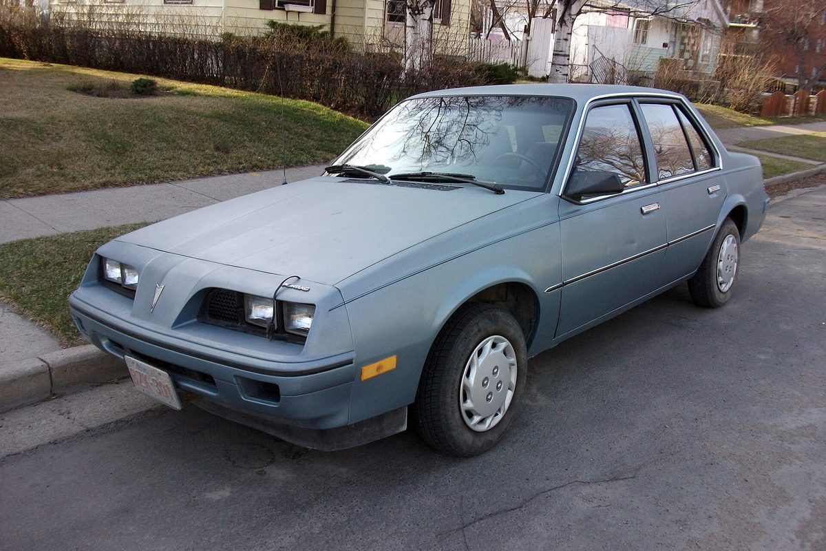 A faded pale blue 1985 Pontiac Sunbird four-door sedan with plain hubcaps, parked at a curb on a residential street