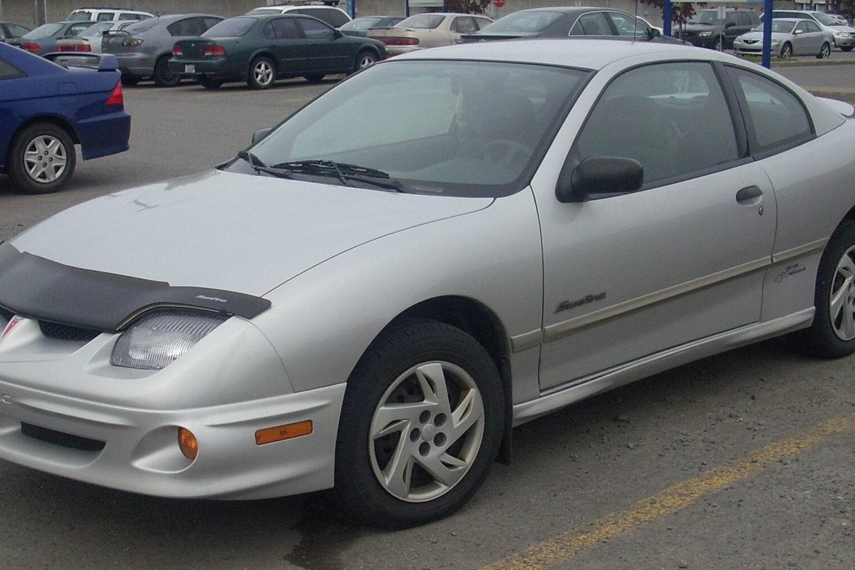 A silver Pontiac Sunfire coupe with a black bug shield on the hood, parked in a busy lot