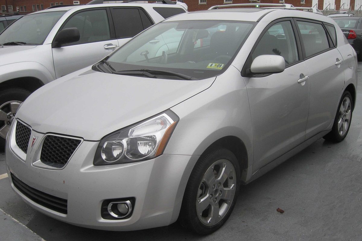 A silver second-generation Pontiac Vibe hatchback with fog lights, parked in a lot between two SUVs