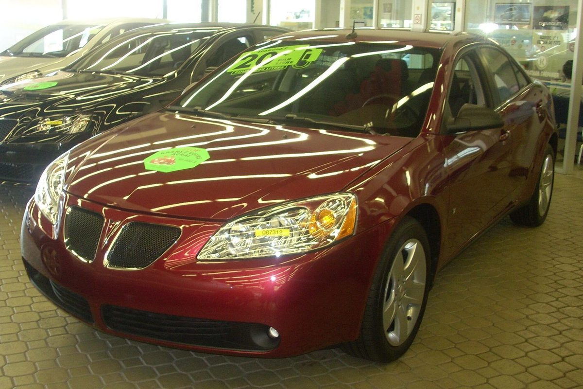 A dark red Pontiac G6 sedan of the 2005-2010 generation parked on a tiled showroom floor, with stickers on its windshield and hood