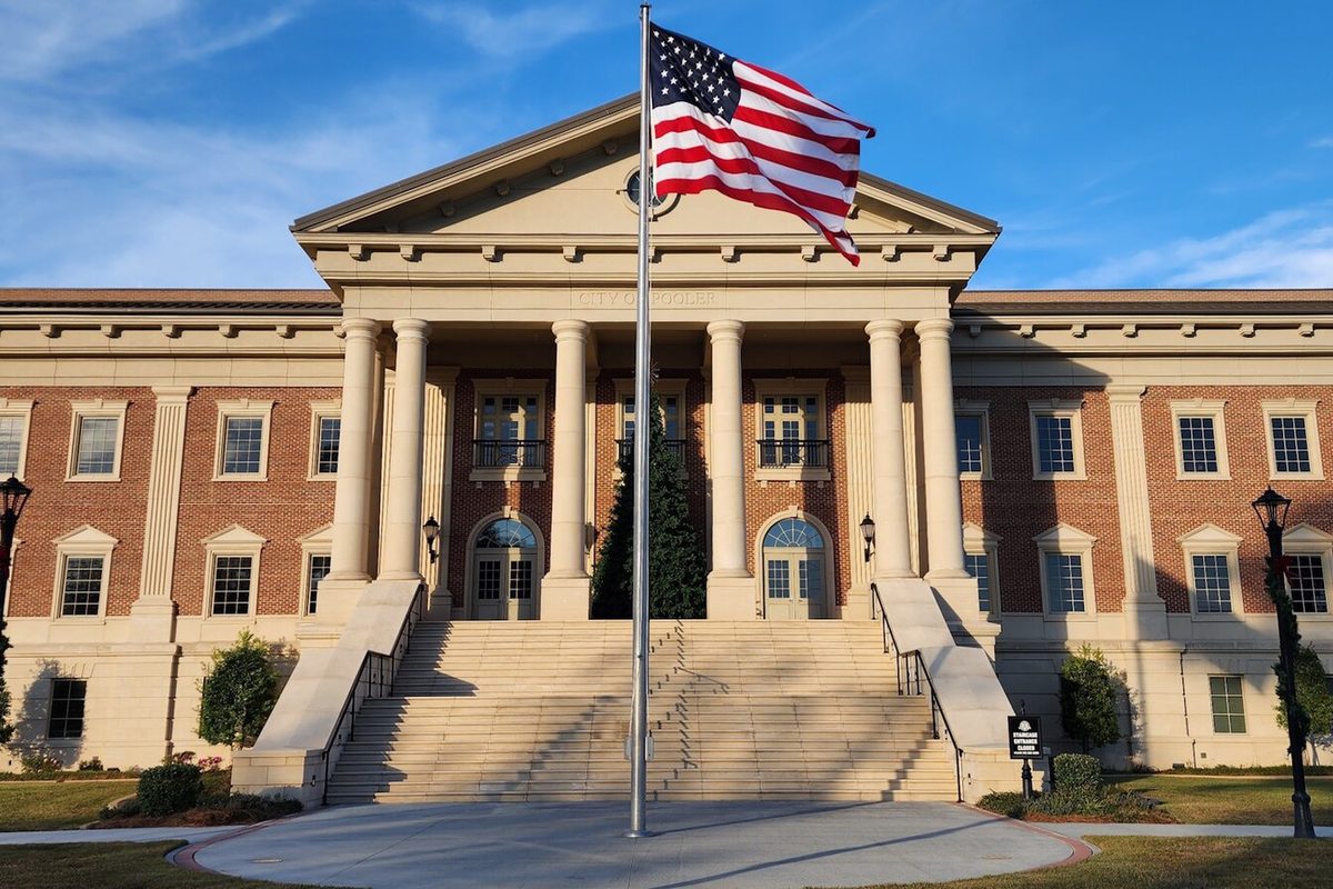 Pooler City Hall, a brick building with a white-columned portico and wide front steps, an American flag flying out front under a blue sky