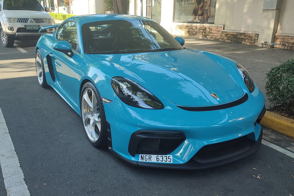 A bright blue Porsche 718 Cayman GT4 with a rear wing and silver wheels parked at a curb