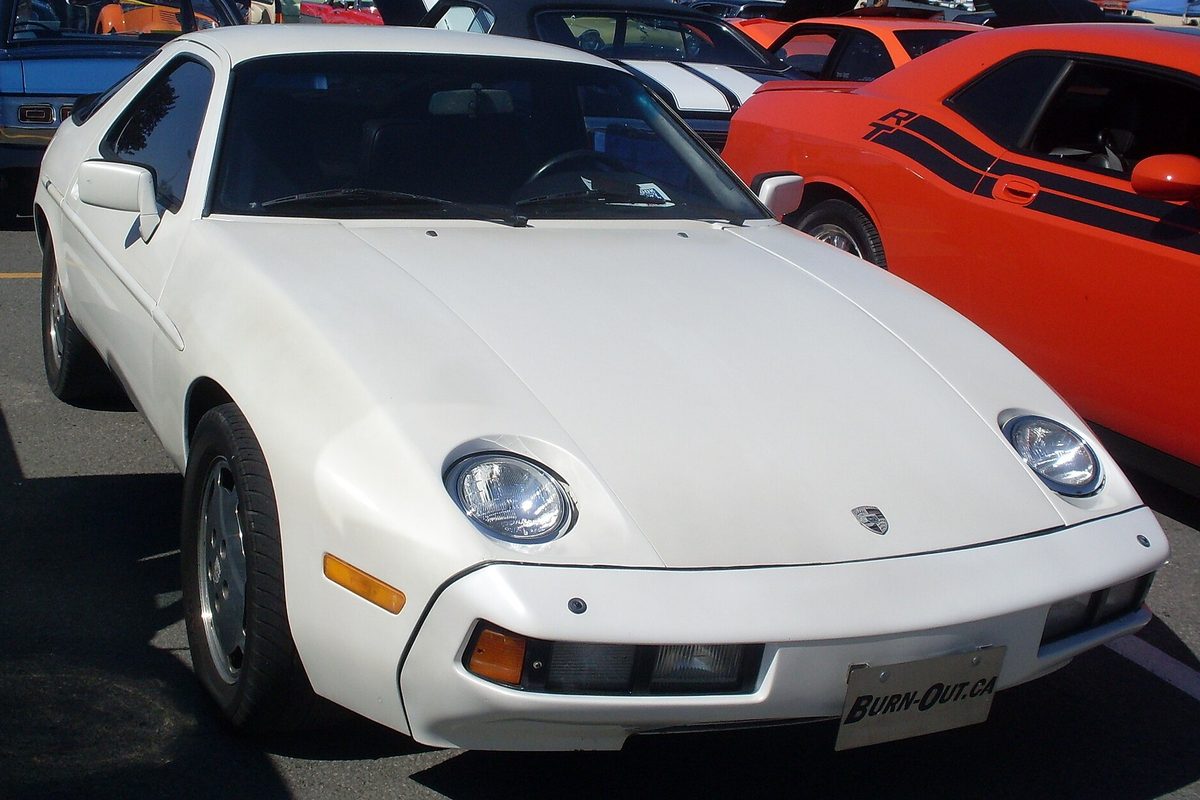 A white Porsche 928 coupe with round exposed headlights parked at an outdoor car meet beside an orange muscle car