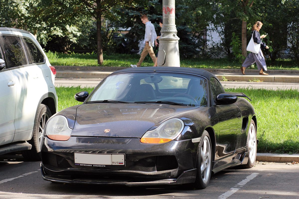 A black Porsche Boxster roadster with its soft top up, parked at a city curb beside a white SUV