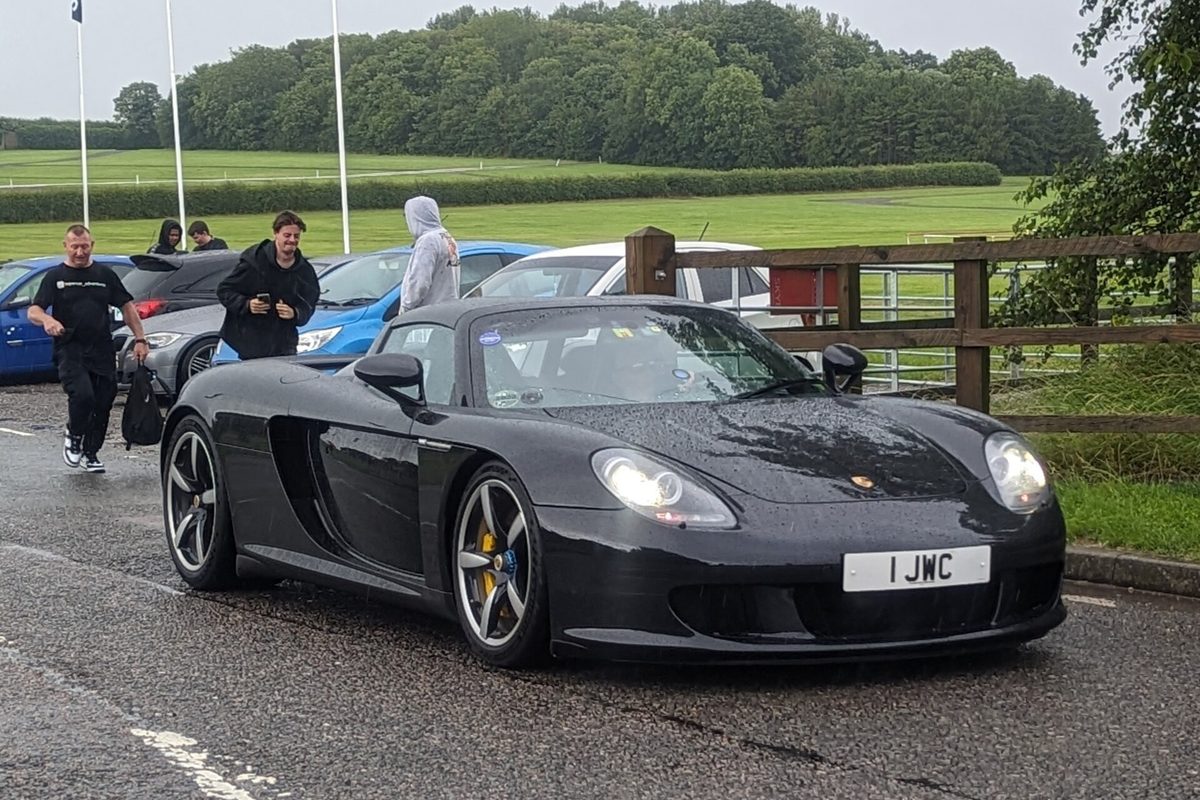 A black Porsche Carrera GT roadster with its headlights on, rolling along a wet road past parked cars and a green field