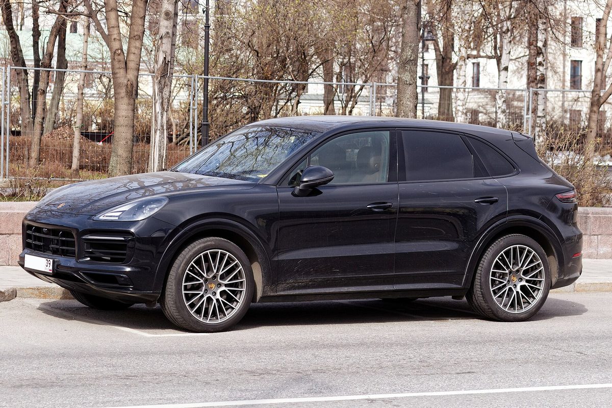 A black Porsche Cayenne SUV with two-tone multi-spoke wheels parked at a city curb, bare trees behind it