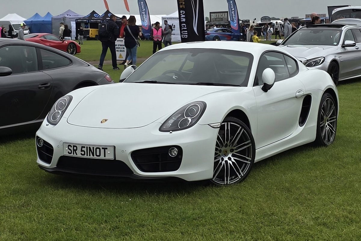 A white Porsche Cayman coupe with dark multi-spoke wheels parked on the grass at an outdoor car show