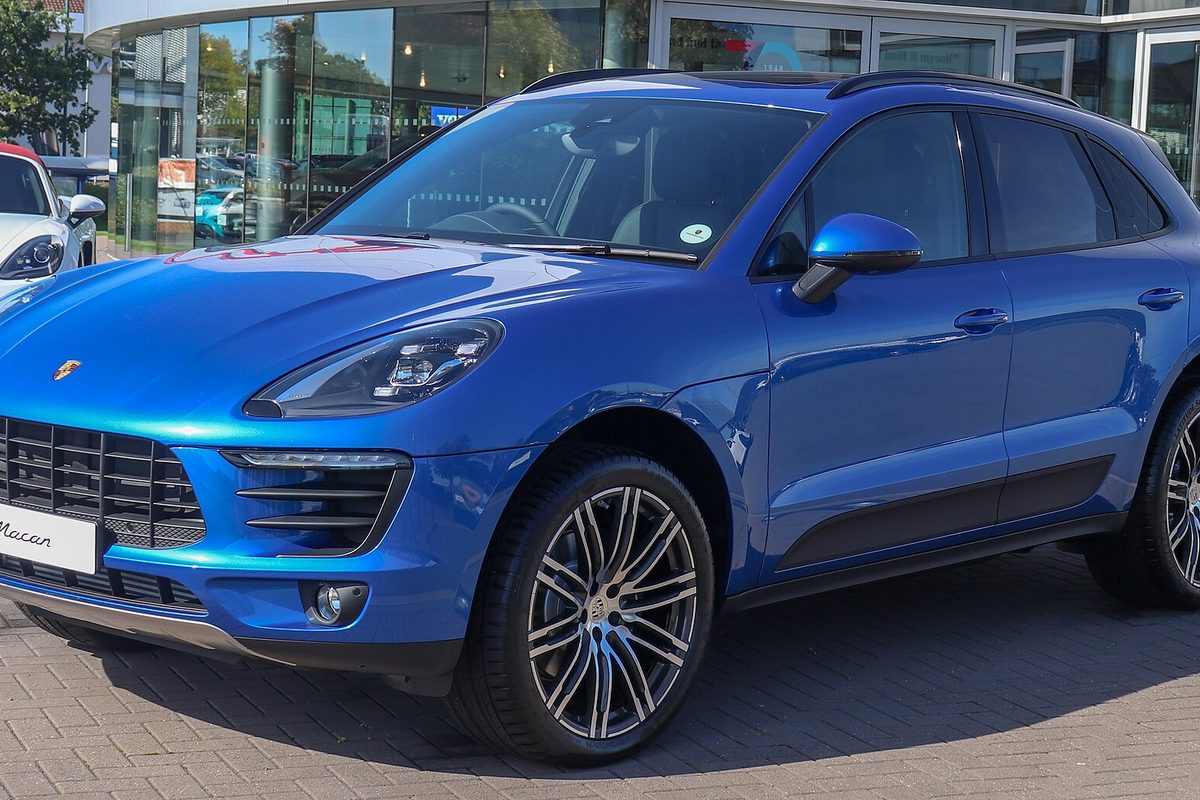 A bright blue Porsche Macan SUV parked on brick paving outside a glass-fronted dealership