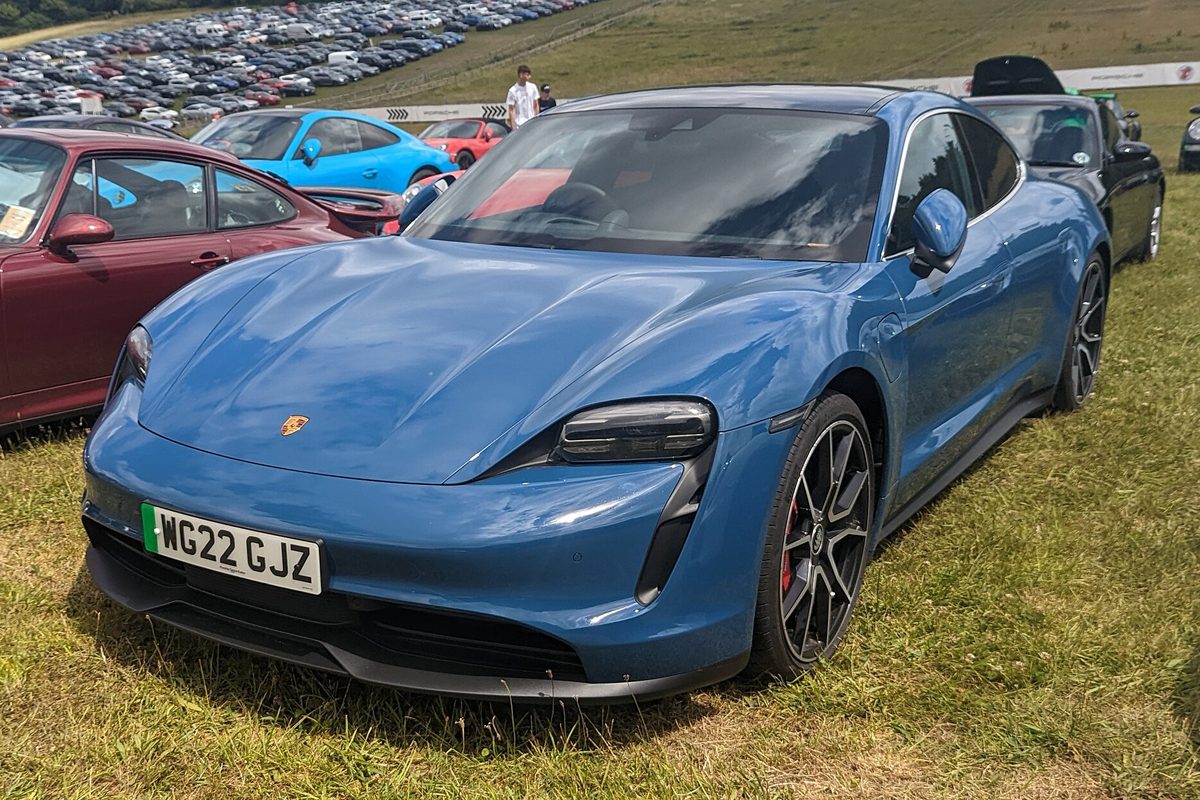 A blue Porsche Taycan sedan parked on grass at a car show, with other Porsches and a full parking field behind it