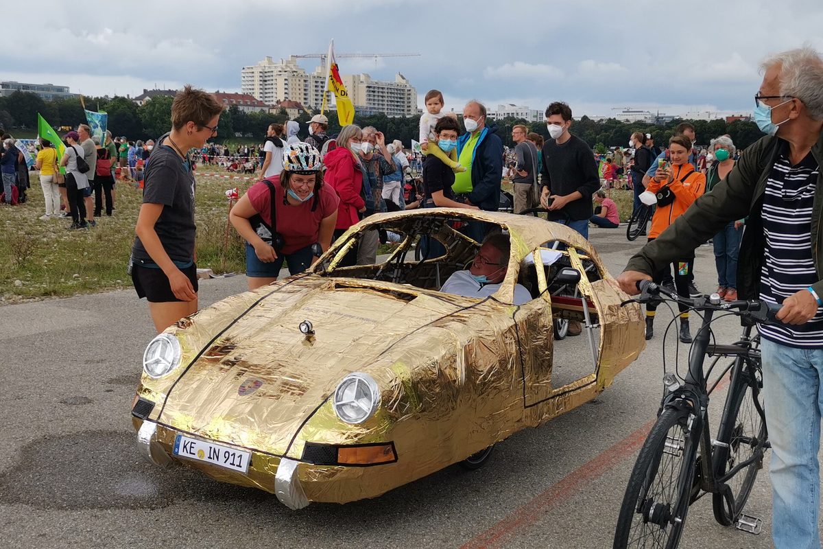 A homemade car wrapped in gold foil and shaped like a classic Porsche, its plate reading KE IN 911, rolling through a crowd at a 2021 transport protest in Munich