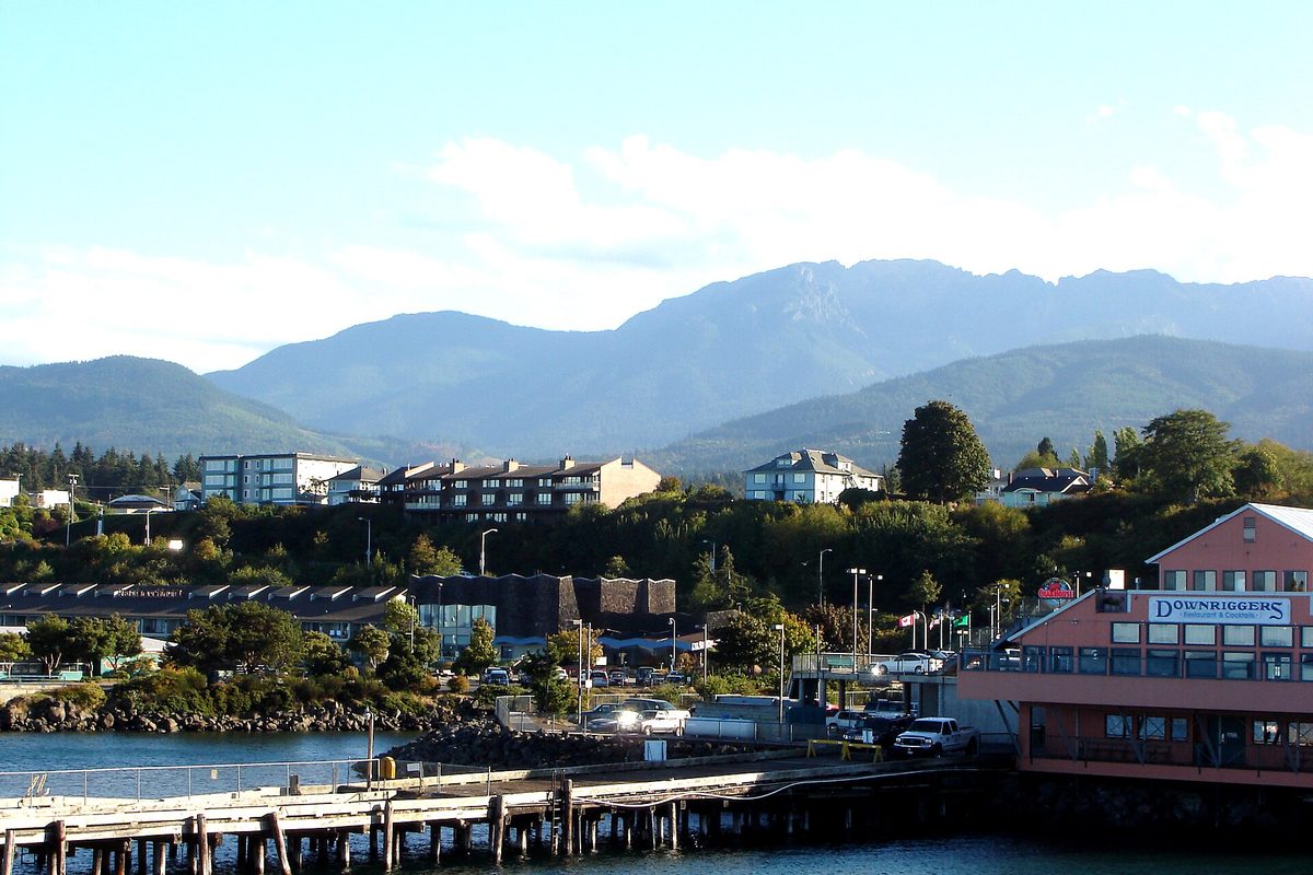 Port Angeles waterfront seen from the ferry, with a wooden pier, a red waterfront building, houses on the hill and the Olympic Mountains behind