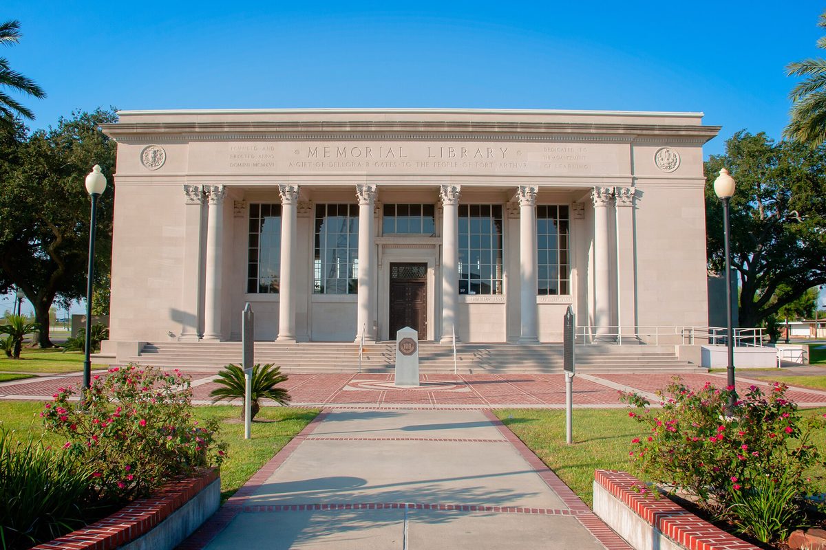 The white columned front of the Gates Memorial Library in downtown Port Arthur, with MEMORIAL LIBRARY carved above the columns, palm trees, a brick walkway and pink flowering shrubs