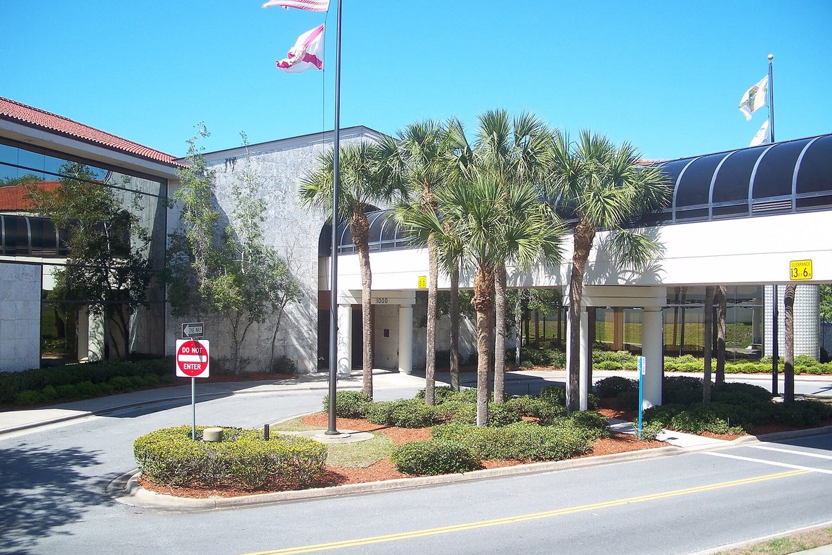 Port Orange City Hall, a white building with a curved covered walkway, flagpoles and a cluster of palm trees on a sunny day