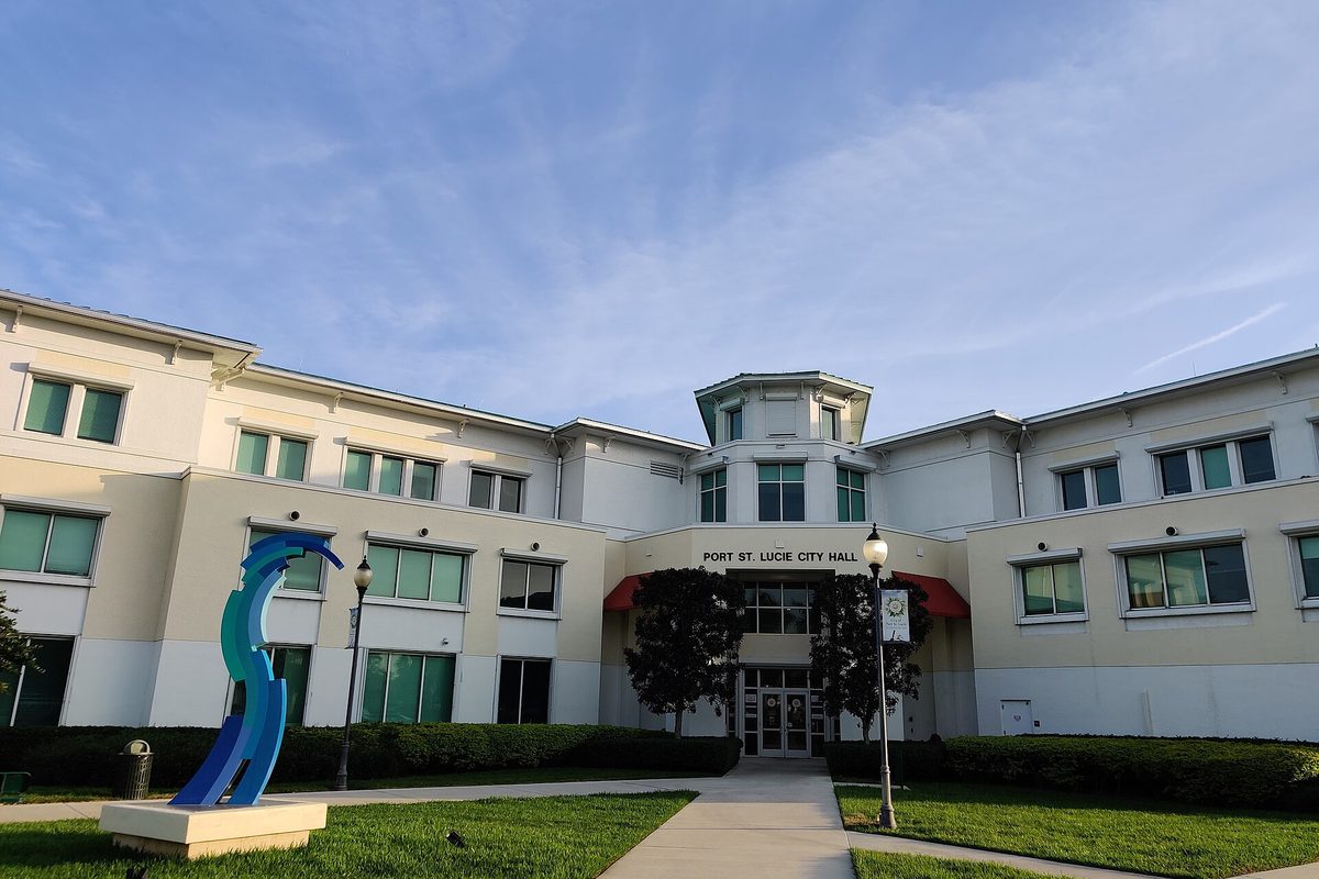 Port St. Lucie City Hall, a cream two-story building with green windows and a red entrance awning, with a blue wave sculpture on the front lawn