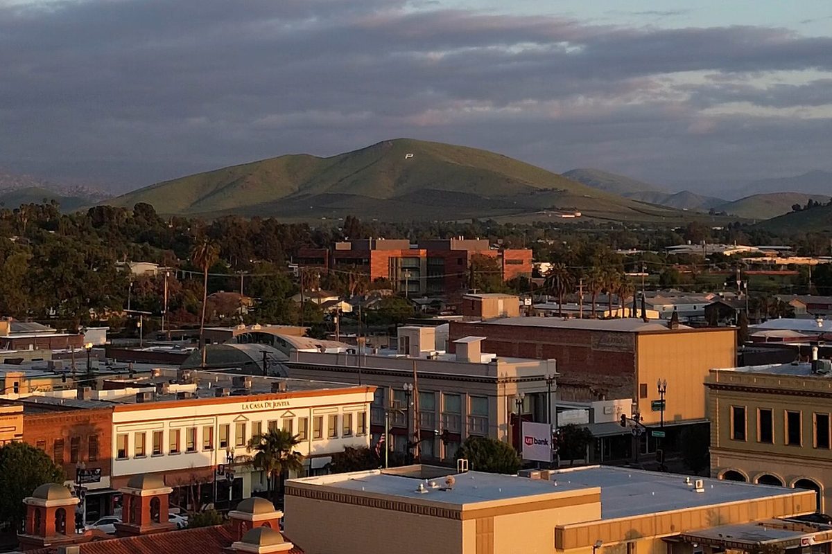 Downtown Porterville's low rooftops and brick buildings at golden hour, with a green hill rising behind the town under heavy clouds