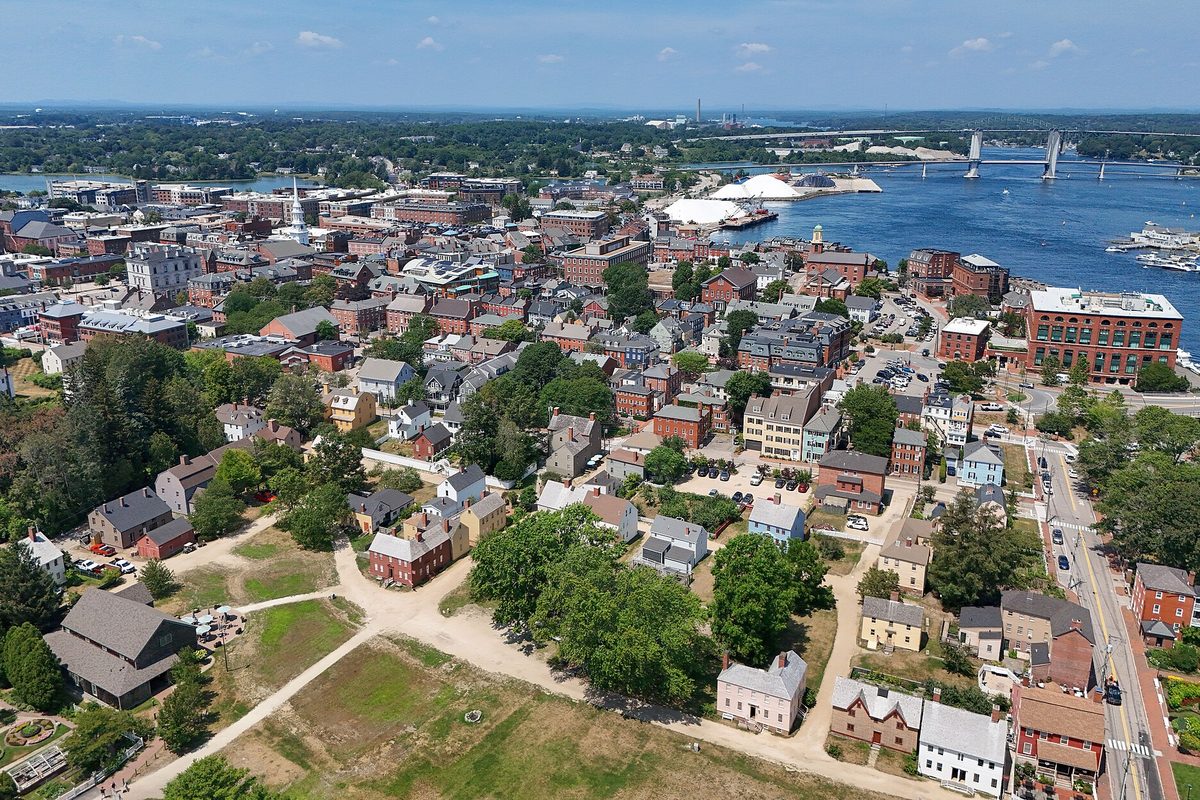 Aerial view of Portsmouth: old clapboard houses around a green lawn in front, brick downtown blocks and a white steeple beyond, and the river with a bridge and harbor in the distance