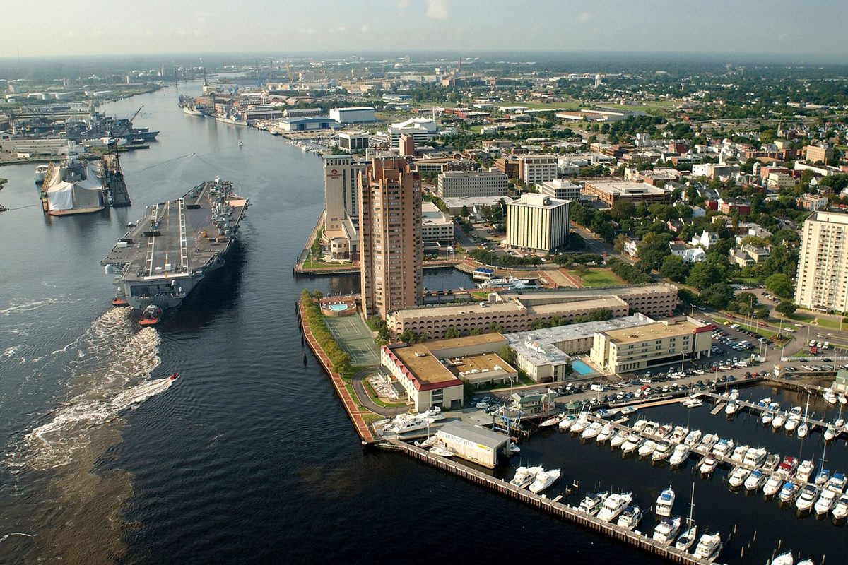 An aircraft carrier moving up the Elizabeth River past the Portsmouth waterfront, with a tall apartment tower, low buildings and a marina full of white boats