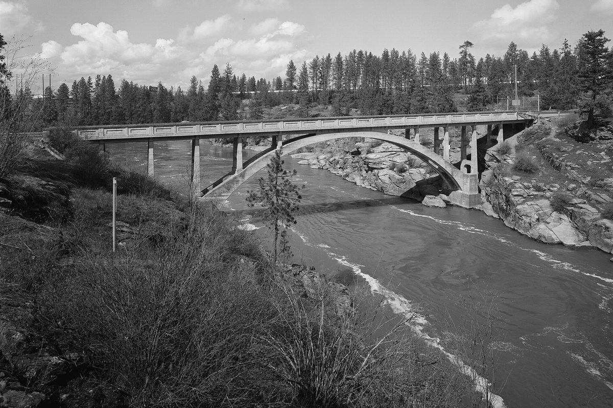 Black-and-white photo of a 1929 concrete arch bridge over the rocky Spokane River at Post Falls, with pine trees on the far bank