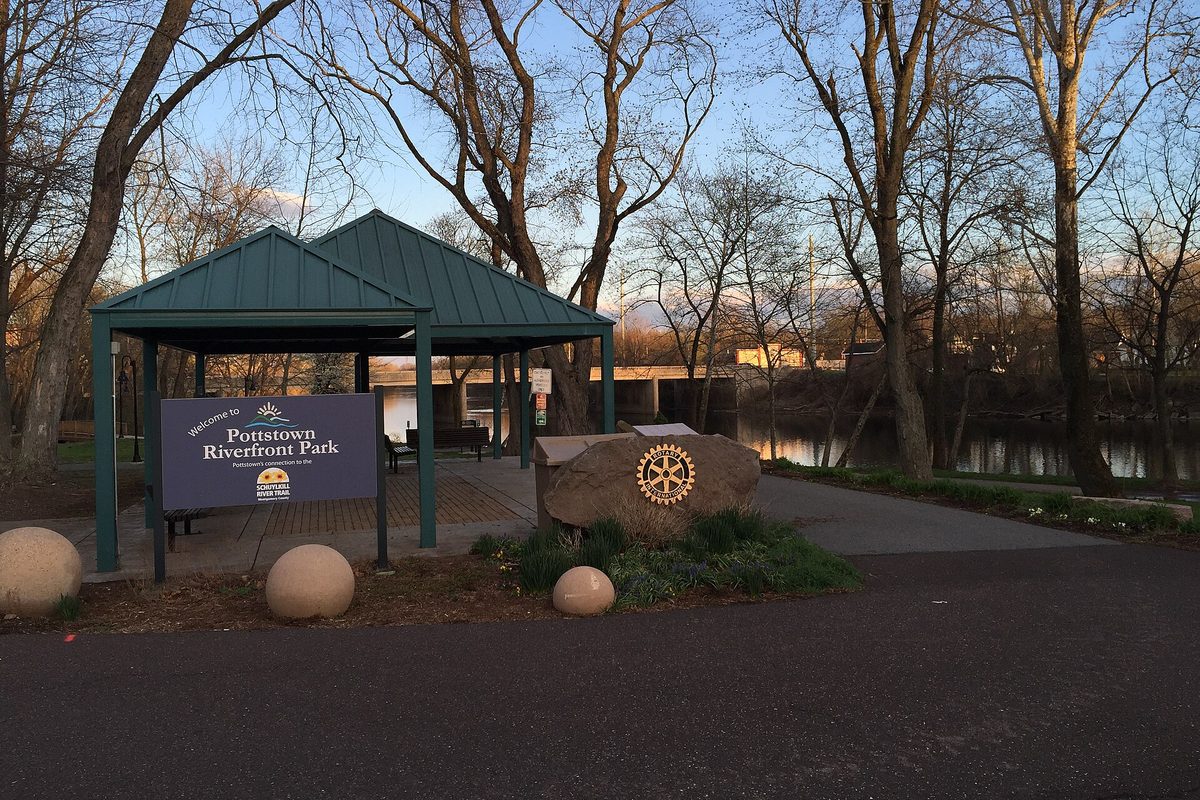 The Pottstown Riverfront Park welcome sign beside a green-roofed pavilion and a stone with a Rotary emblem, with bare trees and still water behind at dusk