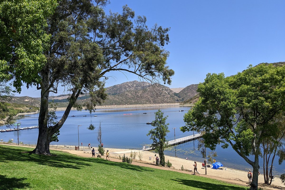 Lake Poway on a sunny day, seen past a large shade tree on a grassy slope, with a sandy shore, floating docks, people walking and dry brown hills across the water