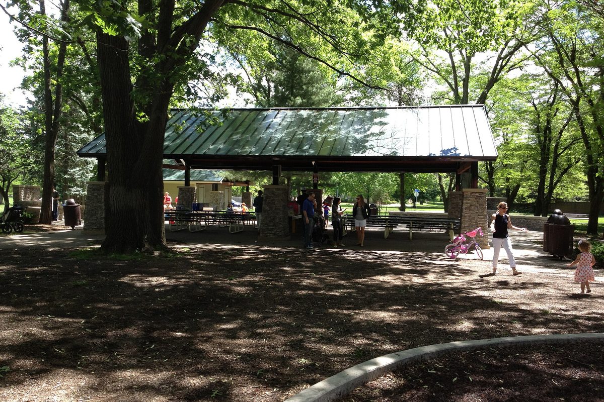 A green-roofed picnic pavilion on stone pillars under tall shade trees in Franklin Park, with families walking nearby