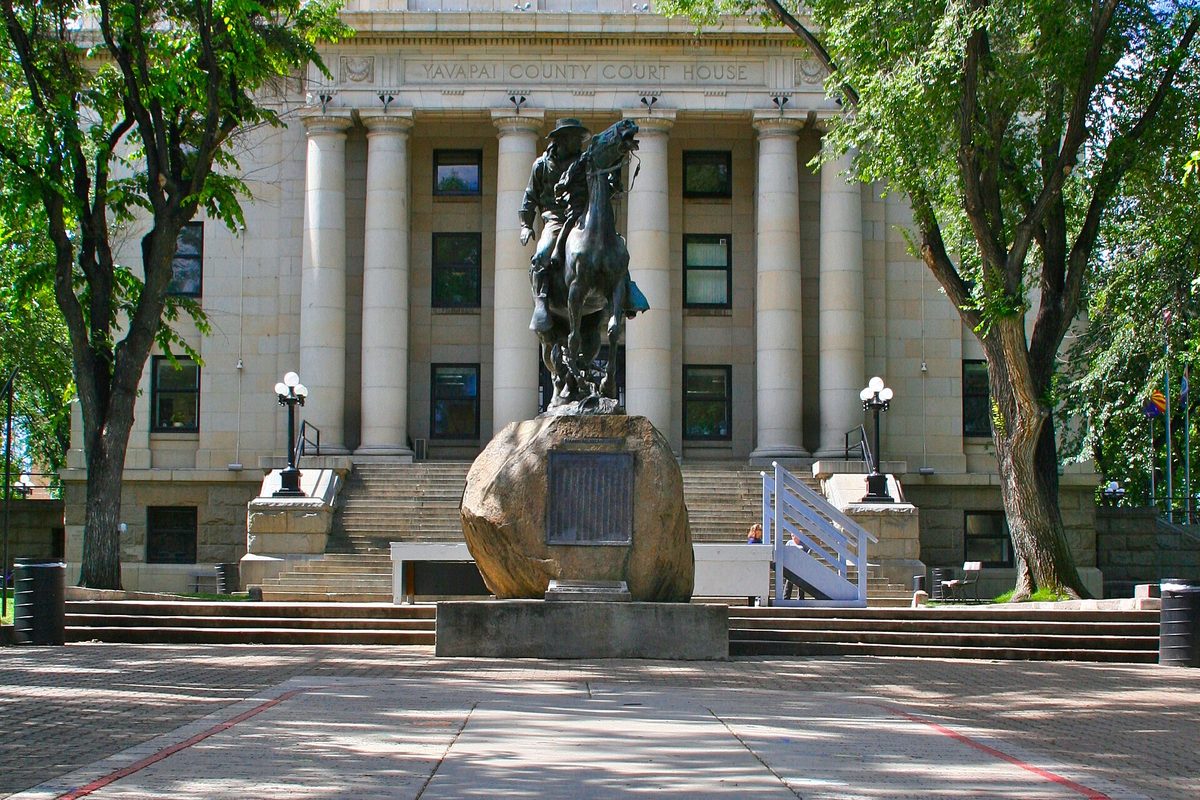 The columned county courthouse in Prescott, with a bronze statue of a rider on horseback on a large boulder in front, framed by green trees