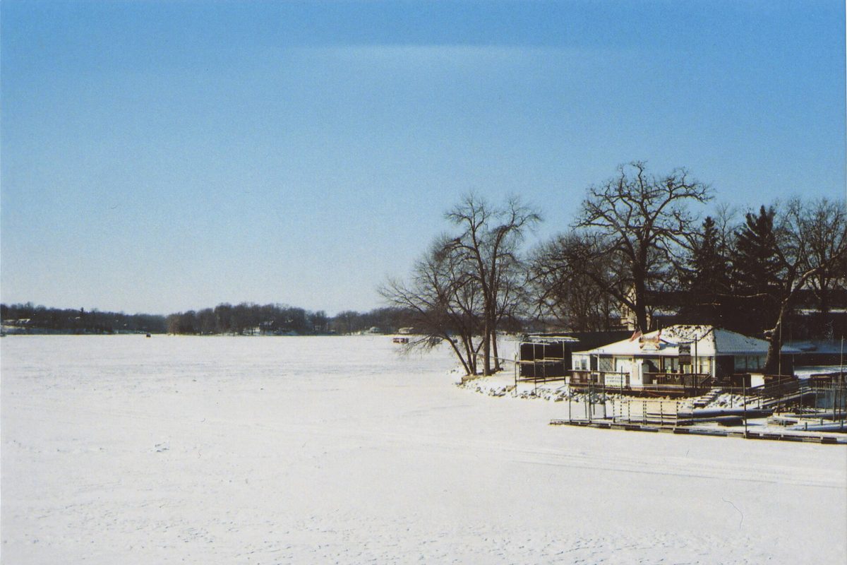 Prior Lake frozen and covered in snow, seen from a bridge, with bare trees and a white lakeside house and docks on the shore under a pale blue sky