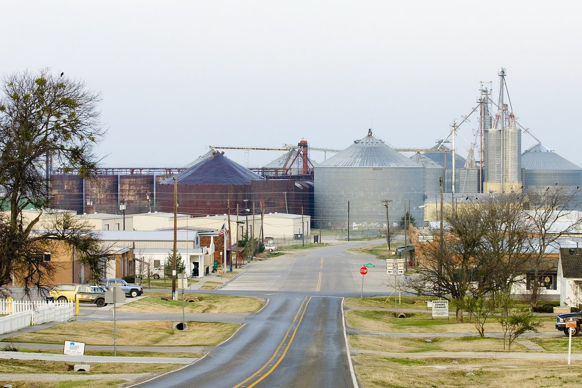 A two-lane street in Prosper running past low buildings and bare trees toward tall grain silos and storage bins under an overcast sky