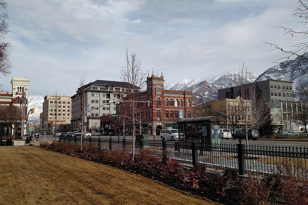 Downtown Provo with a red brick clock tower building between glass-fronted office buildings, snow-covered mountains behind and a lawn and iron fence in front