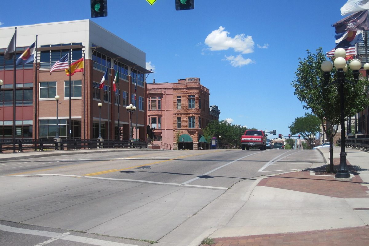 A wide street in downtown Pueblo on a sunny day, with a red pickup heading toward a red brick corner building, a row of flags and globe street lamps