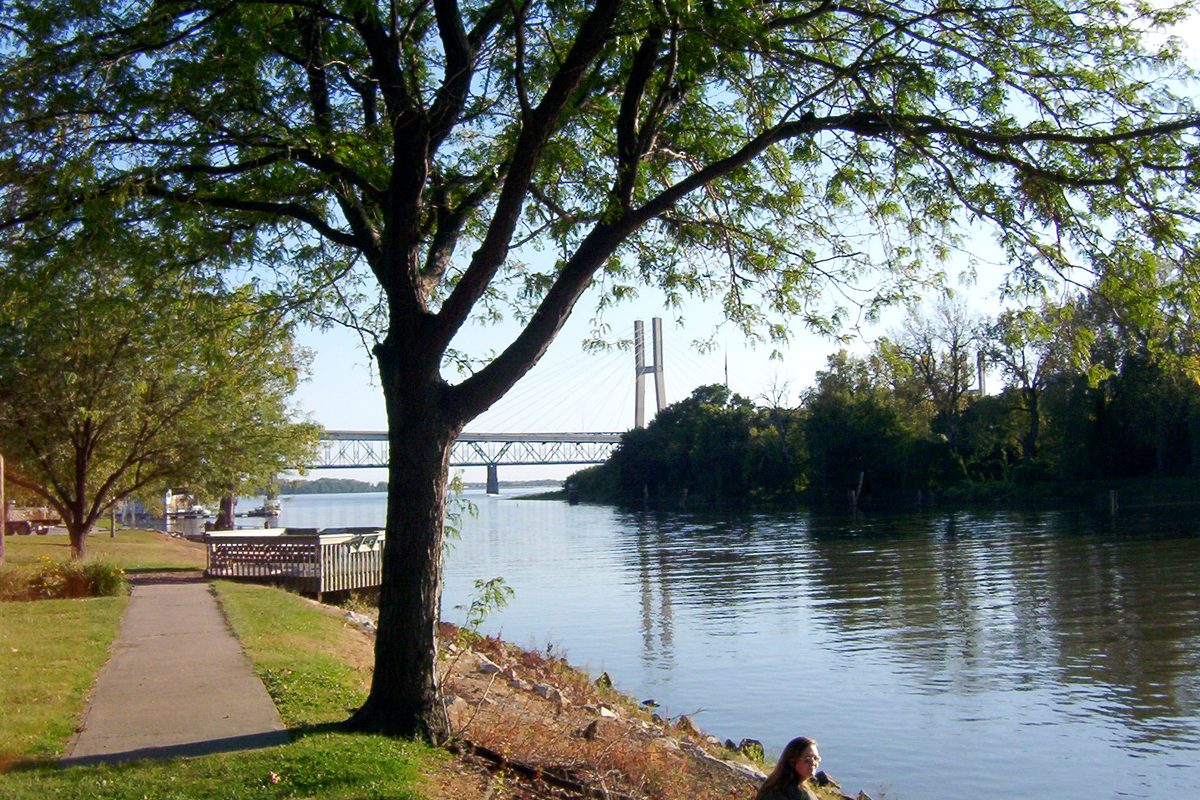 A tree-lined riverfront park path along the Mississippi River in Quincy, with a cable-stayed highway bridge and a truss bridge in the distance