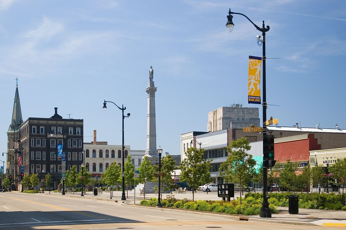 Downtown Racine with a tall stone column monument, street lamps, young trees and brick storefronts under a clear blue sky