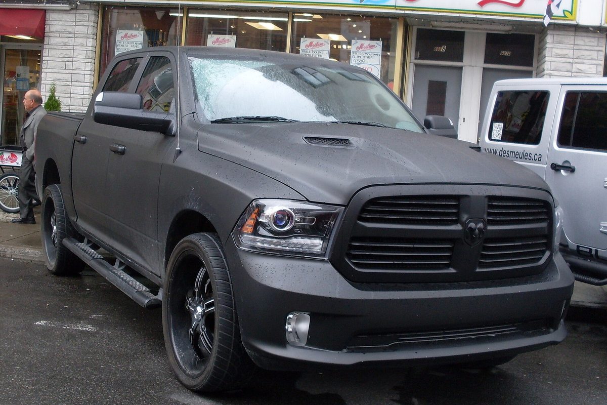 A matte black 2013 Ram 1500 four-door pickup on black wheels, parked on a wet city street in front of a row of shops