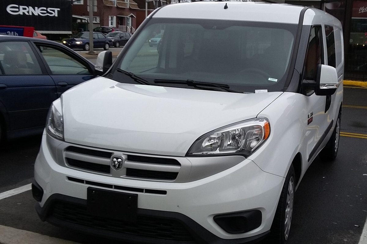 A white Ram ProMaster City compact van seen from the front, parked at a city curb beside a dark sedan