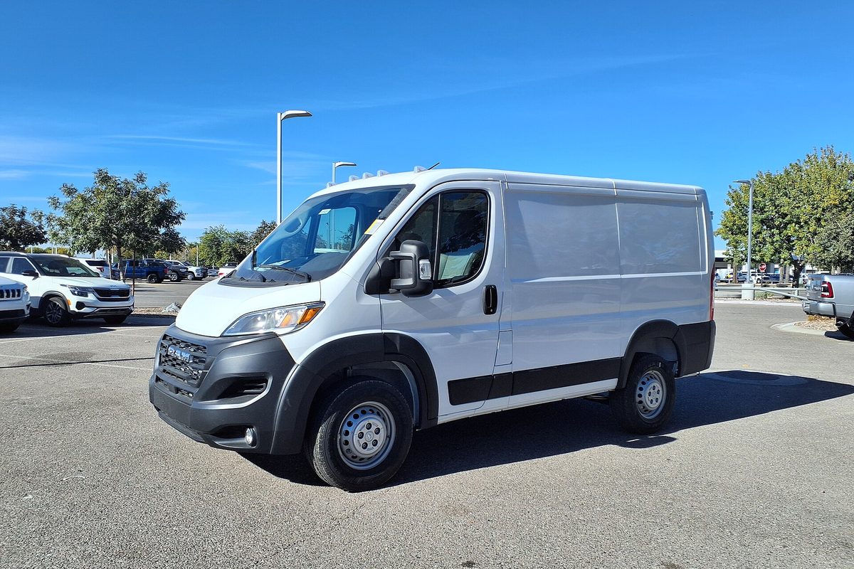 A white low-roof Ram ProMaster cargo van with black bumpers and steel wheels, parked on a sunny dealer lot