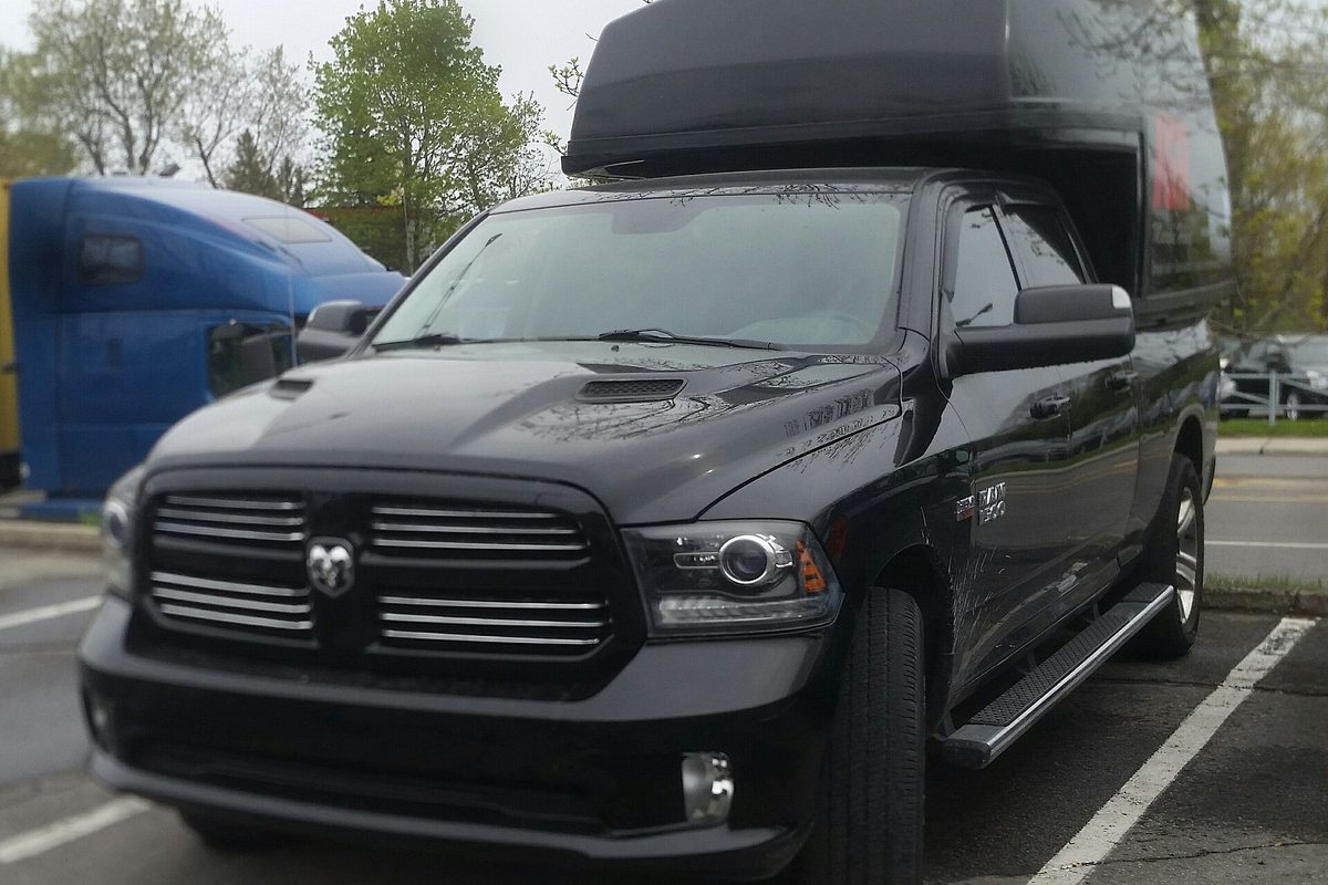 A black Ram 1500 Crew Cab pickup with a tall black cap over its bed, parked in a lot in St. Eustache, Quebec, with a blue semi truck behind it