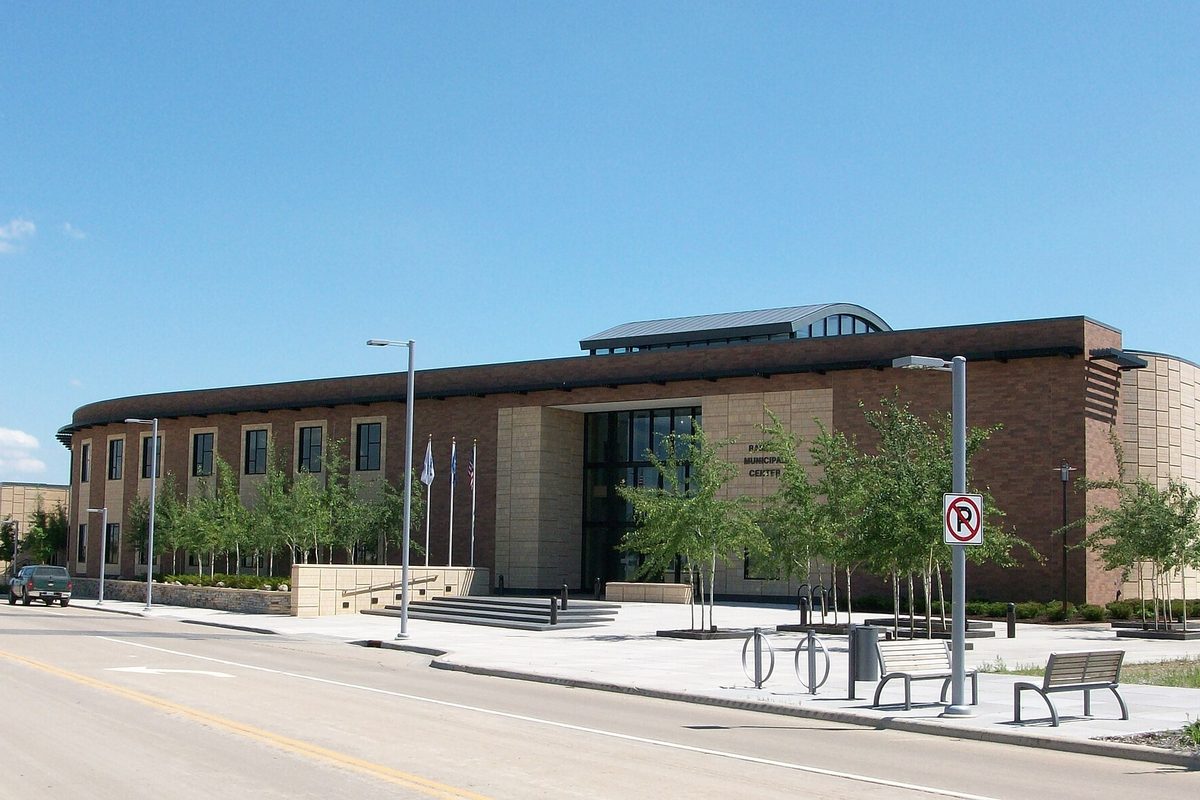 Ramsey Municipal Center on Sunwood Drive, a long two-story brick and tan stone building with a curved roof, young trees, benches and a no-parking sign along the street under a blue sky