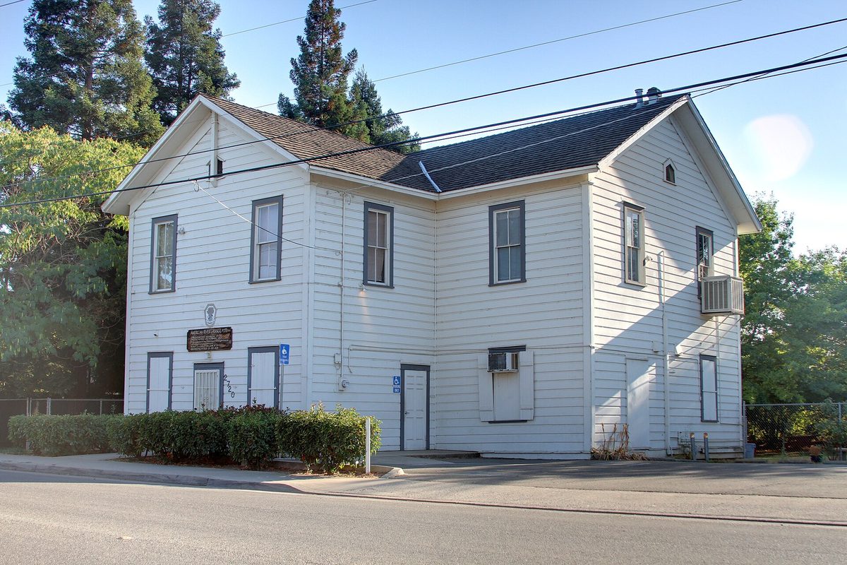 The white two-story wooden American River Grange Hall No. 172 in Rancho Cordova, with tall pines behind it and a sidewalk in front