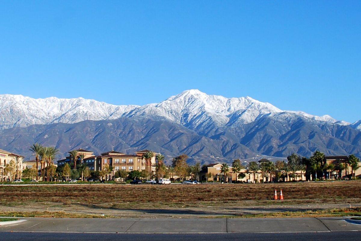 Snow-covered mountain peaks rising behind palm trees and tan apartment buildings in Rancho Cucamonga, with an empty lot and orange cones in front