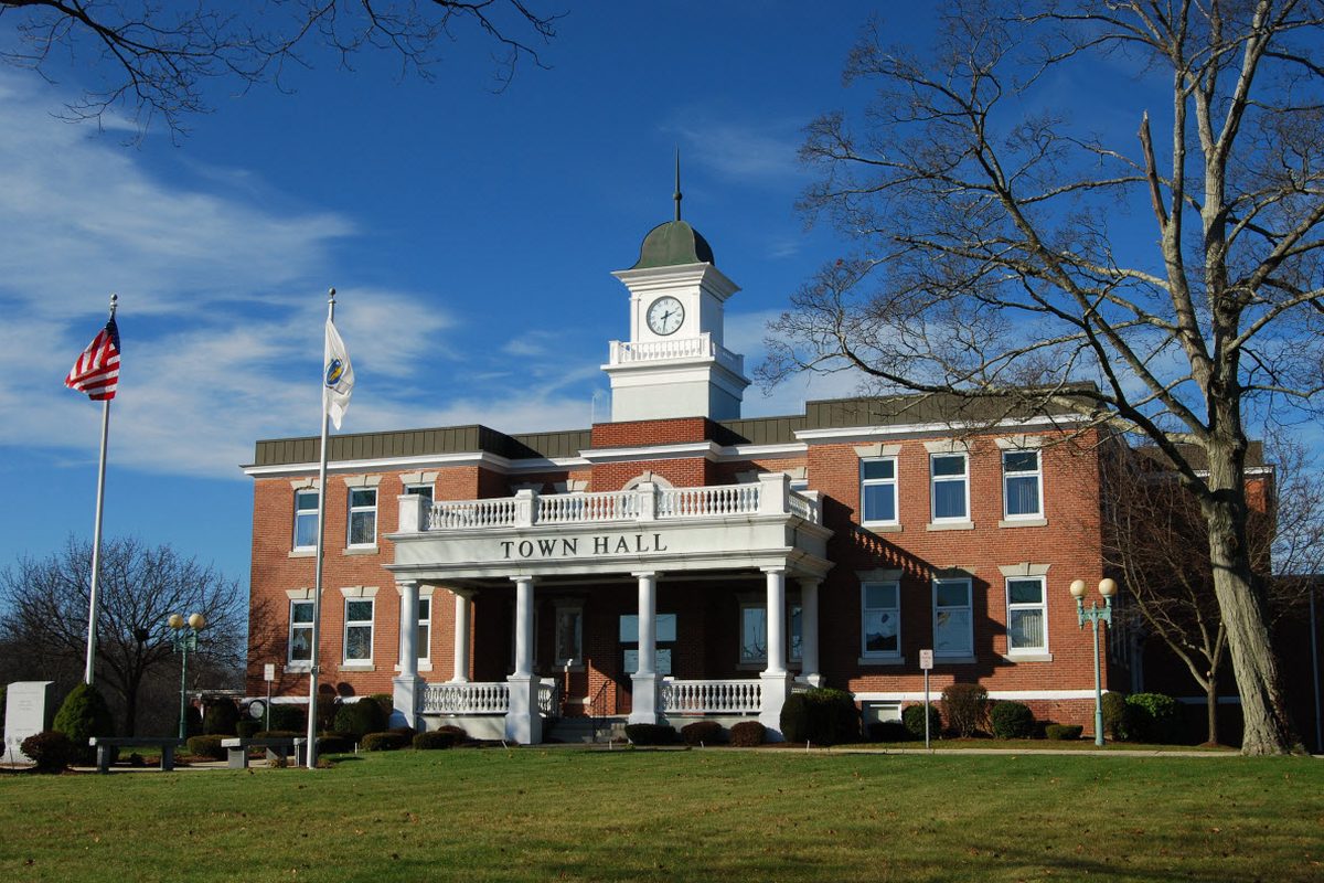 Randolph Town Hall, a red brick building with a white clock tower and a columned front porch, under a blue sky beside a tall bare tree