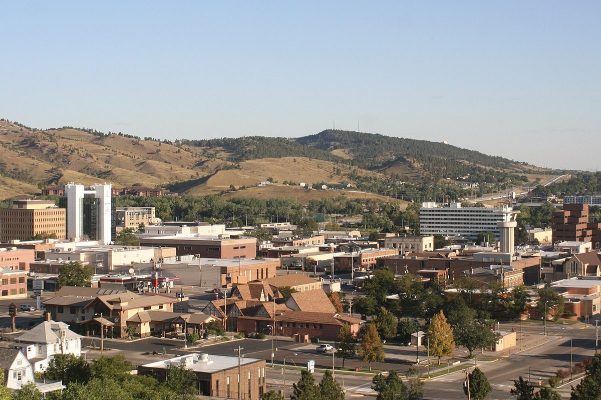 Rapid City seen from Hillcrest Drive: low brick downtown buildings and a few taller towers, with dry, pine-dotted hills behind under a clear sky