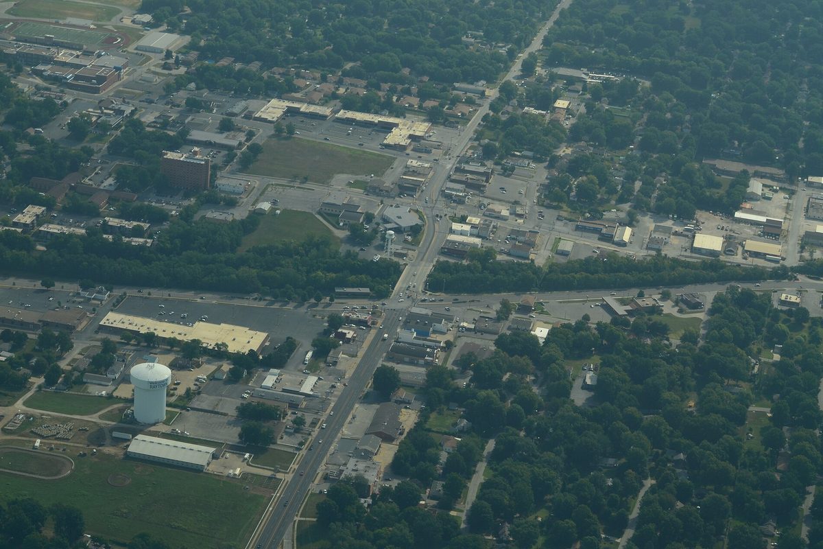 Aerial view of Raytown: a white water tower, a busy crossroads and low commercial roofs set among thick green trees